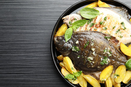 Homemade seafood dish. Tasty roasted flounder fish with herbs and potato on black table, top view....