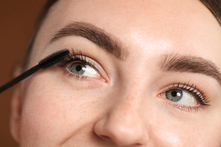 Young woman applying mascara onto her eyelashes on brown background, closeup