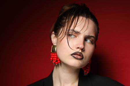Beautiful young woman with black lip liner wearing stylish earrings on dark red background