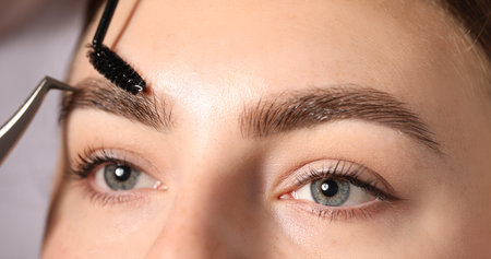 Plucking and brushing young woman's eyebrow with spoolie on light grey background, closeup