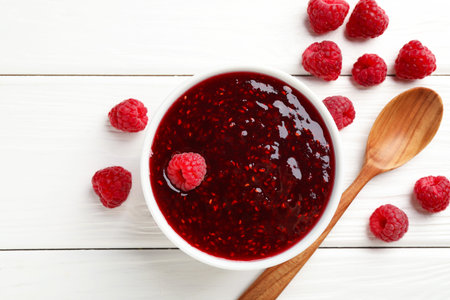 Sweet raspberry jam in bowl and berries on white wooden table, flat lay
