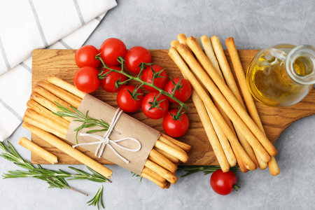 Delicious grissini sticks, rosemary, oil and fresh tomatoes on gray textured table, flat lay