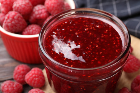 Sweet raspberry jam in glass jar and berries on table, closeup