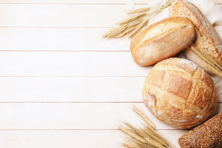Different types of bread and wheat spikes on wooden table, flat lay. Space for text