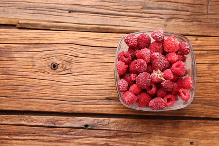 Ripe frozen raspberries in bowl on wooden table, top view. Space for text