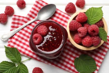Tasty jam, fresh ripe raspberries and leaves on white wooden table, flat lay.