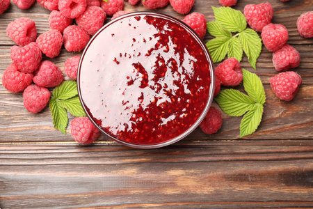 Delicious raspberry jam in glass bowl, fresh berries and mint on wooden table, flat lay