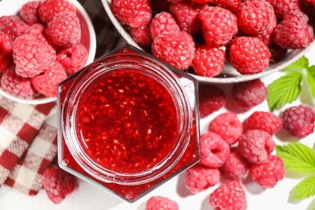 Delicious raspberry jam in glass jar, fresh berries, mint and checkered napkin on white table, flat...