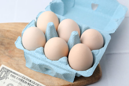 Raw chicken eggs in egg carton and dollar banknote on white tiled table, closeup