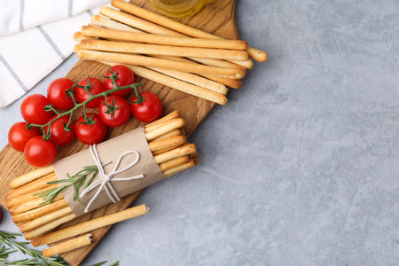 Delicious grissini sticks, rosemary and fresh tomatoes on grey textured table, flat lay. Space for...