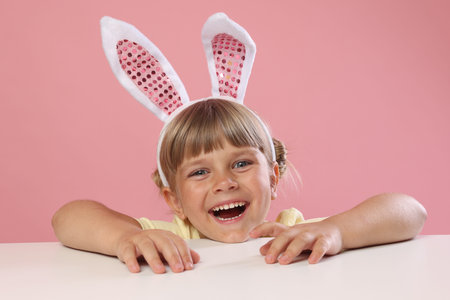 Curious little girl in bunny ears peeking over white table against pink background