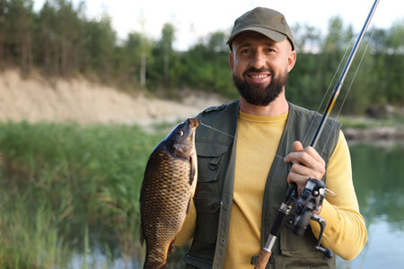 Smiling fisherman holding rod and caught fish near lake at summer