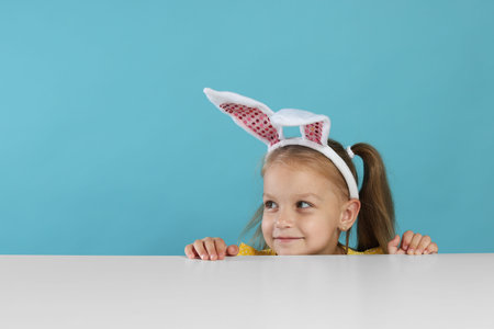 Curious little girl in bunny ears peeking over white table against light blue background, space for...