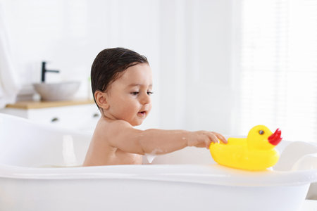 Cute little baby with rubber duck bathing in tub at home