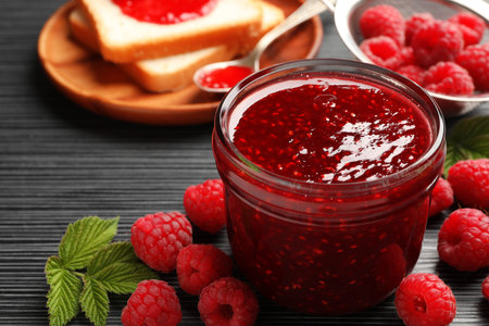 Sweet raspberry jam in glass jar, berries and bread slices on dark gray wooden table, closeup