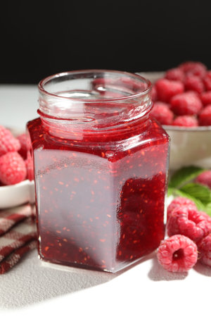 Delicious raspberry jam in glass jar, fresh berries and mint on white textured table against black...