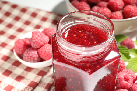 Delicious raspberry jam in glass jar, fresh berries, mint and checkered napkin on white table,...