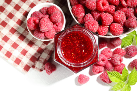 Delicious raspberry jam in glass jar, fresh berries, mint and checkered napkin on white table, flat...