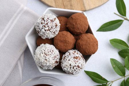 Homemade candies with cocoa powder and coconut flakes in bowl on white table, flat lay