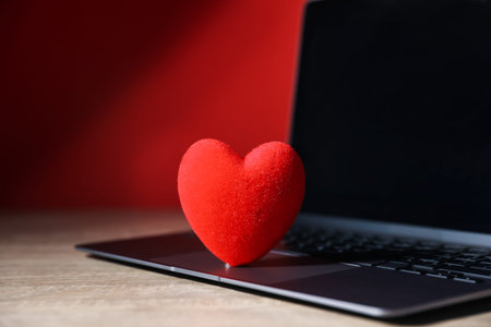 Online dating site. Red heart and laptop on wooden table, closeup