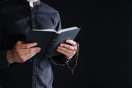 Priest with rosary and Bible on black background, closeup. Space for text