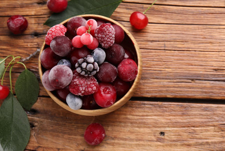 Mix of ripe frozen berries in bowl and green leaves on wooden table, flat lay. Space for textの写真素材