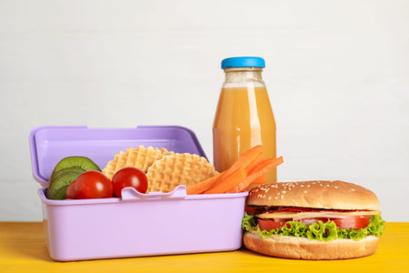 Open lunch box with delicious food and juice on yellow wooden table against light grey background,...