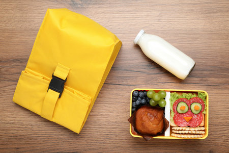 Open lunch box with tasty food, bottle of milk and bag on wooden table, flat lay. Back to school