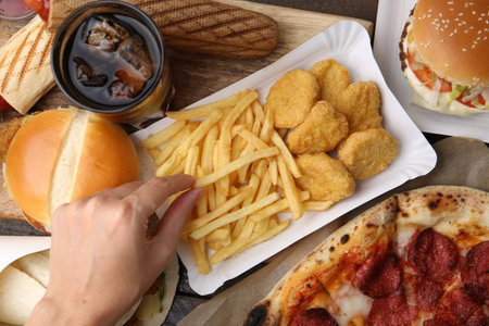Woman taking tasty French fry at wooden table with different fast food and cola, top view