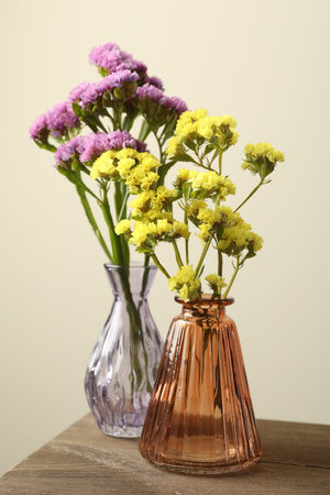 Beautiful flowers in glass vases on wooden table against beige background, closeup