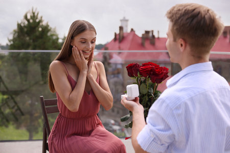 Man with engagement ring and bouquet of roses making marriage proposal to his happy girlfriend on balcony outdoorsの写真素材