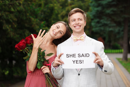 Smiling man holding sheet of paper with text She Said Yes and his happy beloved with bouquet of roses showing ring on engagement day outdoorsの写真素材