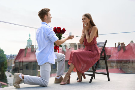 Man with engagement ring and bouquet of roses making marriage proposal to his happy girlfriend on balcony outdoors, low angle viewの写真素材