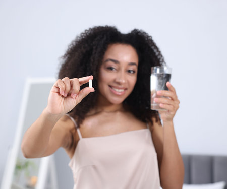 Beautiful Brazilian woman with glass of water and collagen pill indoors, selective focusの写真素材
