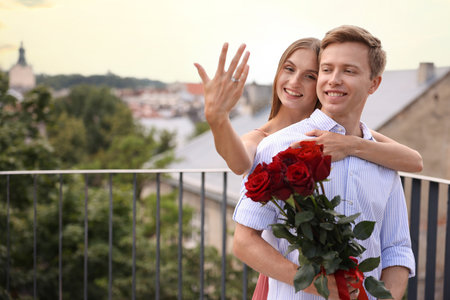 Marriage proposal. Smiling woman showing engagement ring and her fiance holding bouquet of roses on balcony outdoors. Space for textの写真素材