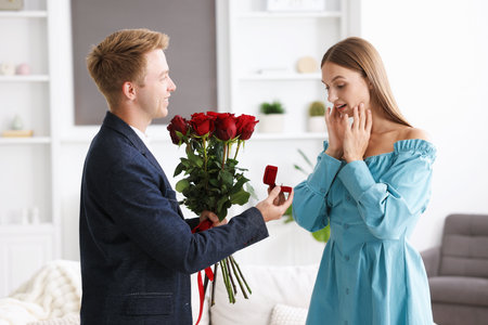 Man with engagement ring and bouquet of roses making marriage proposal to his happy girlfriend at homeの写真素材