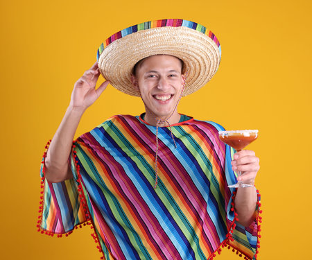 Happy man in sombrero hat, colorful poncho and cocktail on yellow backgroundの写真素材