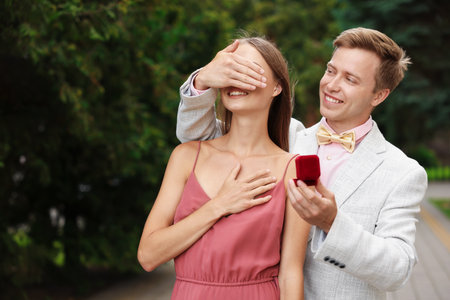 Making marriage proposal. Smiling man with engagement ring surprising his girlfriend outdoors, space for textの写真素材