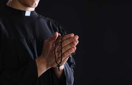 Priest with rosary praying on black background, closeup. Space for textの写真素材