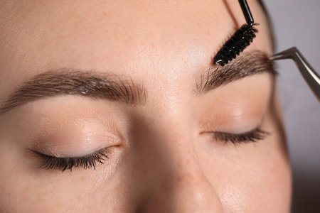 Plucking and brushing young woman's eyebrow with spoolie on light grey background, closeupの写真素材