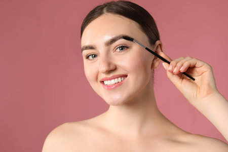 Young woman brushing her eyebrow with spoolie on pink background