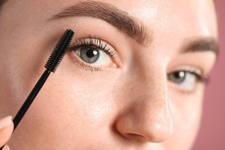 Young woman applying mascara onto her eyelashes on pink background, closeupの写真素材