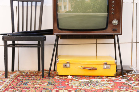 Retro TV on stand, suitcase and black chair near white wall.