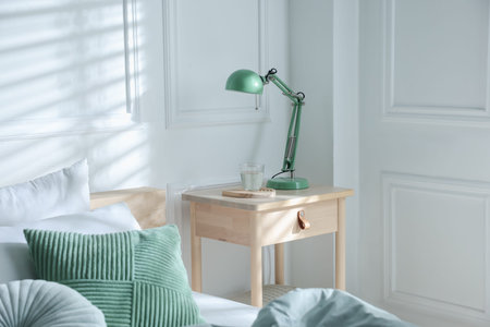 Bedside table with lamp and glass of water near bed indoors