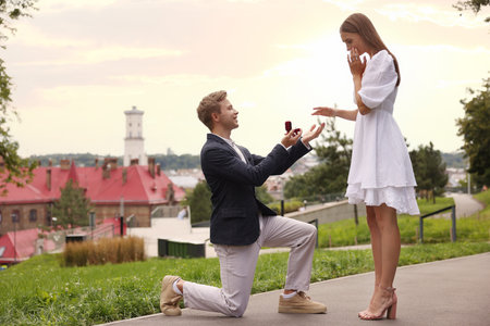 Smiling man with engagement ring making marriage proposal to his surprised girlfriend outdoors....
