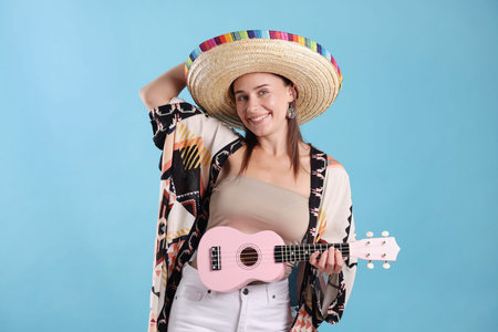 Happy woman in sombrero hat playing ukulele on light blue background