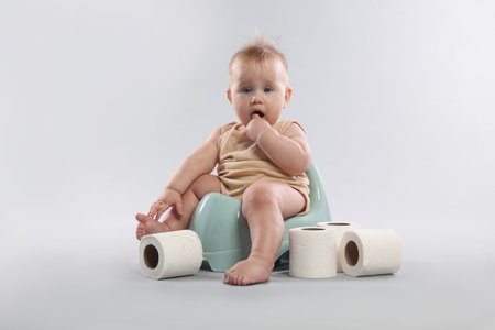 Cute little baby sitting on potty and rolls of toilet paper against light grey background