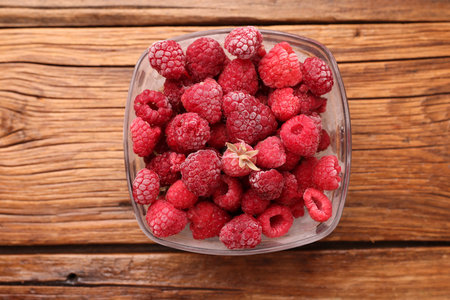 Ripe frozen raspberries in bowl on wooden table, top view
