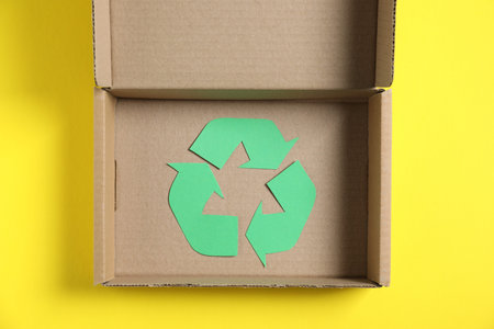 Eco friendly packaging. Recycling symbol and cardboard box on yellow background, top view