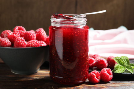 Raspberry jam in glass jar, spoon, fresh berries and mint on wooden table, closeup